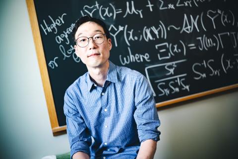 Taeho Kim wearing a blue button-up shirt and glass sitting in front of a black board with math problems on it.